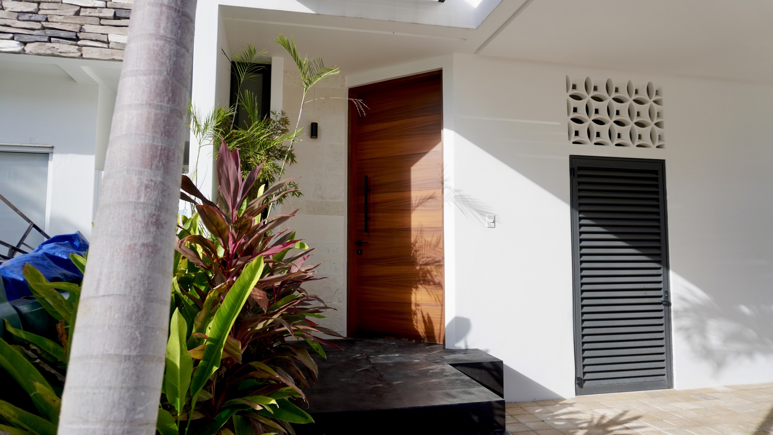 Casa en venta av huayacan - Entrada principal con puerta de madera, jardín tropical y diseño minimalista en fachada blanca